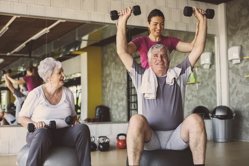 Twee oudere deelnemers trainen met handgewichten onder begeleiding van een trainer